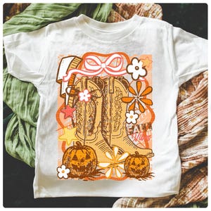 May include: White t-shirt featuring a retro-style graphic with cowboy boots, pumpkins, and flowers in shades of orange, brown, and white. The text "Fall Vibes" is also included in the design. The shirt is made of a soft material.