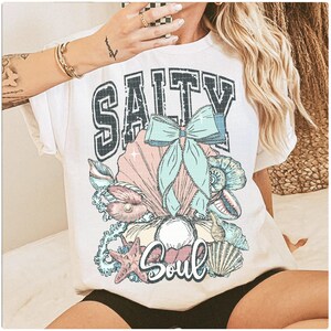 Puede incluir: Camiseta blanca con un estampado gr&aacute;fico de conchas marinas, un lazo y el texto "Salty Soul".