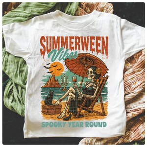 May include: White t-shirt with a colorful graphic featuring a skeleton relaxing on a beach chair under an umbrella, holding a glass of wine. The text reads "SUMMERWEEN VIBES" and "SPOOKY YEAR ROUND".