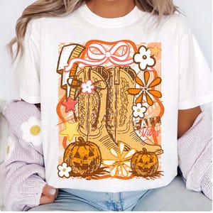 May include: White t-shirt featuring a graphic design with cowboy boots, pumpkins, and flowers in shades of orange and brown. The words "Fall Vibes" are also included in the design.