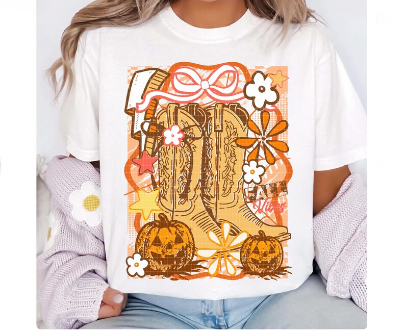 May include: White t-shirt featuring a graphic design with cowboy boots, pumpkins, and flowers in shades of orange and brown. The words "Fall Vibes" are also included in the design.