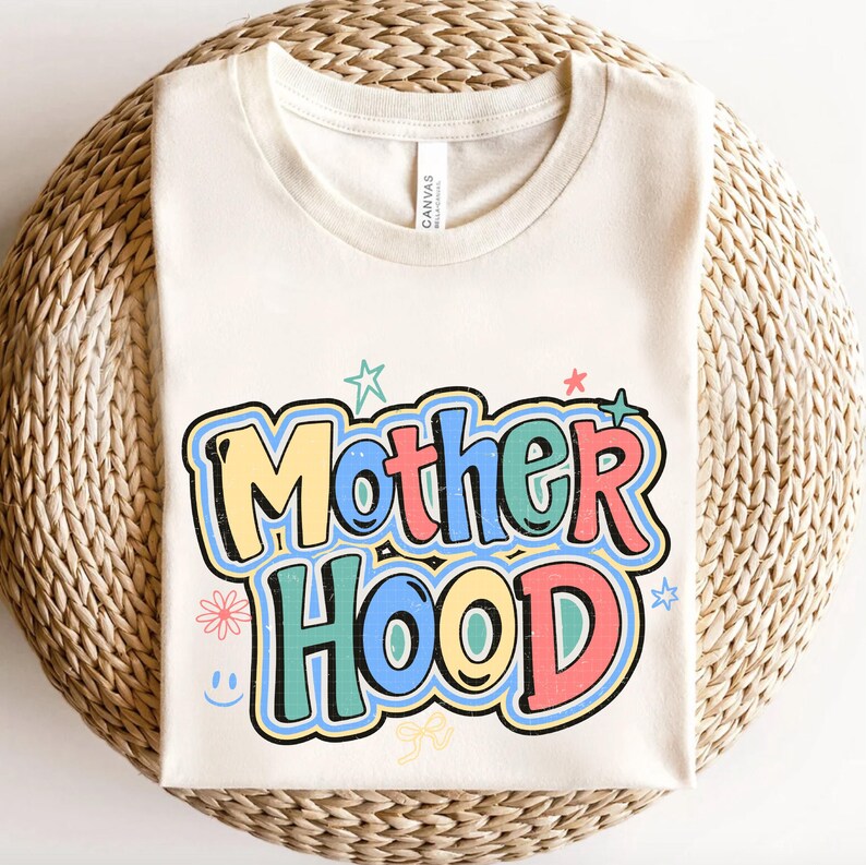 May include: A cream-coloured t-shirt featuring the word "Motherhood" in a vibrant, retro font. The design incorporates stars, flowers, and a smiley face. The shirt is made of a soft material and is placed on a woven surface.