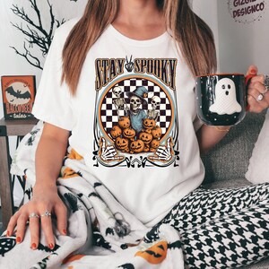 Puede incluir: Camiseta blanca con un dise&ntilde;o circular de cuadros blancos y negros. El c&iacute;rculo presenta un esqueleto con un sombrero de bruja y sosteniendo una calabaza. El texto "Stay Spooky" est&aacute; por encima del c&iacute;rculo.
