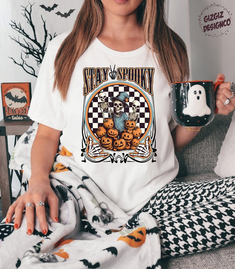 Puede incluir: Camiseta blanca con un dise&ntilde;o circular de cuadros blancos y negros. El c&iacute;rculo presenta un esqueleto con un sombrero de bruja y sosteniendo una calabaza. El texto "Stay Spooky" est&aacute; por encima del c&iacute;rculo.