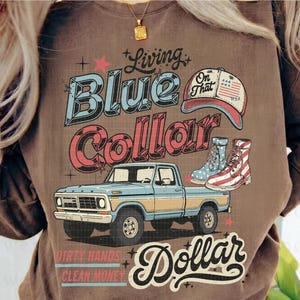 May include: Brown t-shirt with a graphic design featuring a blue and yellow pickup truck, a baseball cap, and boots. The text reads "Living Blue Collar Dollar" with additional text "Dirty Hands Clean Money."