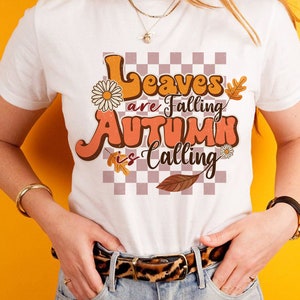 Op de afbeelding: Wit T-shirt met een retro-grafische afbeelding met de tekst "Leaves are Falling Autumn is Calling". De afbeelding heeft een geruite achtergrond, herfstbladeren en bloemen.