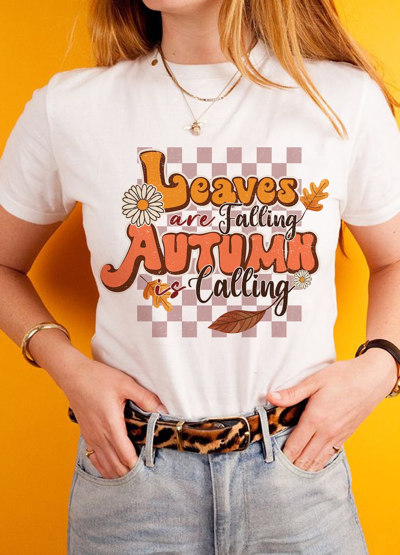 Op de afbeelding: Wit T-shirt met een retro-grafische afbeelding met de tekst "Leaves are Falling Autumn is Calling". De afbeelding heeft een geruite achtergrond, herfstbladeren en bloemen.