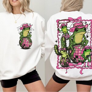 May include: White sweatshirt featuring a cartoon frog wearing a pink cowboy hat, sitting on a pink disco ball. The back of the sweatshirt has a larger graphic of frogs, cactus, and a lasso, all in a pink frame.