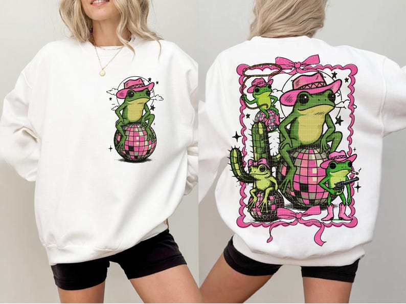 May include: White sweatshirt featuring a cartoon frog wearing a pink cowboy hat, sitting on a pink disco ball. The back of the sweatshirt has a larger graphic of frogs, cactus, and a lasso, all in a pink frame.