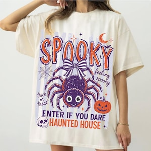 May include: An off-white t-shirt featuring a graphic print with the text SPOOKY, feeling spooky, trick or treat, ENTER IF YOU DARE, and HAUNTED HOUSE. The design includes a large spider with a bow, a pumpkin, a spiderweb, a crescent moon, and a skull.