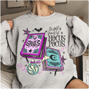 May include: A light grey sweatshirt with a Halloween-themed design. The graphic features spell books, a potion bottle, stars, and a bat. The text on the sweatshirt reads "It's just a bunch of Hocus Pocus" and "Spells".
