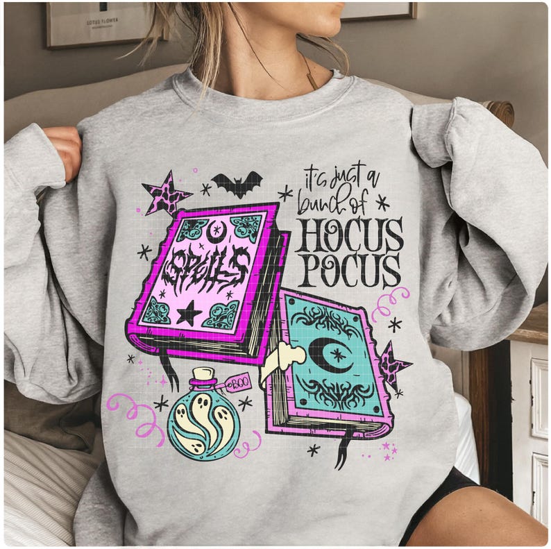 May include: A light grey sweatshirt with a Halloween-themed design. The graphic features spell books, a potion bottle, stars, and a bat. The text on the sweatshirt reads "It's just a bunch of Hocus Pocus" and "Spells".