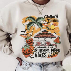 May include: Cream-colored sweatshirt with a graphic design featuring a palm tree, beach umbrella, and jack-o'-lantern strawberries. The text reads "Chillin' & Thrillin' SUMMER-WEEN VIBES". The design has orange, black, and green accents.