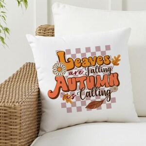 Op de afbeelding: Een wit kussen met een geruite achtergrond en de tekst "Leaves are falling Autumn is calling" in oranje en bruine letters. Het kussen heeft een bloemenmotief en een blad.