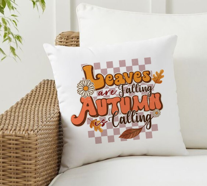 Op de afbeelding: Een wit kussen met een geruite achtergrond en de tekst "Leaves are falling Autumn is calling" in oranje en bruine letters. Het kussen heeft een bloemenmotief en een blad.