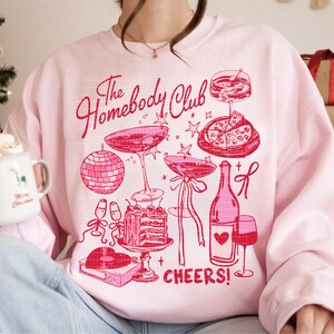 May include: Pink sweatshirt with a graphic design of a disco ball, drinks, cake, pizza, and the text "The Homebody Club Cheers!"