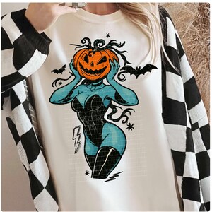 May include: Cream-colored sweatshirt featuring a Halloween-themed graphic. The design depicts a blue figure with a pumpkin head, wearing a black corset and boots. Bats and lightning bolts are also included in the design. The sleeves are black and white checkered.
