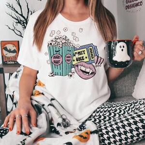 May include: A white t-shirt with a graphic design of a bucket of popcorn with skulls, a vampire mouth, and a movie ticket that says "Admit One" and "Murder".