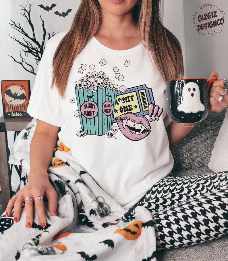 May include: A white t-shirt with a graphic design of a bucket of popcorn with skulls, a vampire mouth, and a movie ticket that says "Admit One" and "Murder".