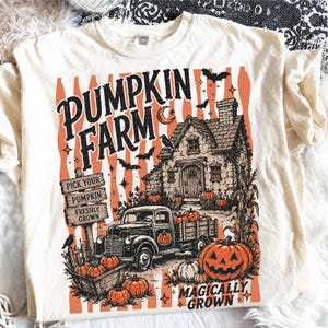 Peut inclure: Un t-shirt cr&egrave;me avec une impression graphique portant le texte PUMPKIN FARM et MAGICALLY GROWN. Le motif comprend un camion vintage, une maison, des citrouilles, des chauves-souris, un corbeau et un panneau en bois PICK YOUR PUMPKIN FRESHLY GROWN.