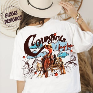 May include: White t-shirt with a vintage-style graphic featuring a cowgirl riding a horse, a barn, and the words "Cowgirls Long Live." The design includes red stars and a windmill.
