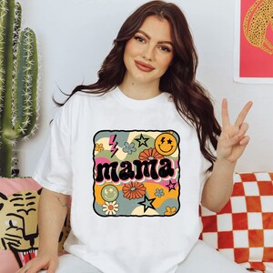Op de afbeelding: Wit T-shirt met een retro-grafisch ontwerp met het woord "mama" in een kleurrijk, golvend lettertype omringd door sterren, bloemen en smiley-gezichten.