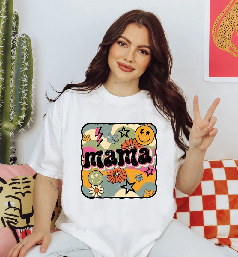Op de afbeelding: Wit T-shirt met een retro-grafisch ontwerp met het woord "mama" in een kleurrijk, golvend lettertype omringd door sterren, bloemen en smiley-gezichten.