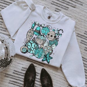 Peut inclure: Sweat-shirt blanc avec un motif d'ornements de No&euml;l bleus et bruns. Les ornements sont dessin&eacute;s dans un style vintage.
