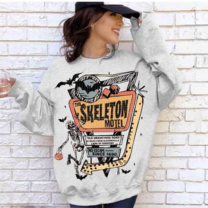 Puede incluir: Una sudadera gris claro con un gr&aacute;fico colorido del letrero "The Skeleton Motel" con un esqueleto, murci&eacute;lagos y texto. El dise&ntilde;o incluye el texto "Old Graveyard Road", "No Escape Since 1690".