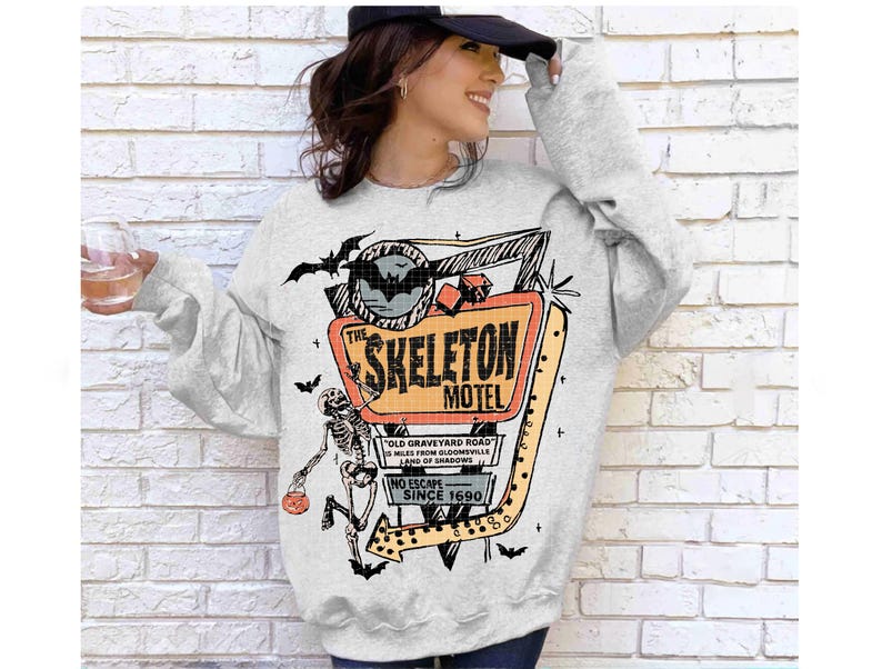 Puede incluir: Una sudadera gris claro con un gr&aacute;fico colorido del letrero "The Skeleton Motel" con un esqueleto, murci&eacute;lagos y texto. El dise&ntilde;o incluye el texto "Old Graveyard Road", "No Escape Since 1690".