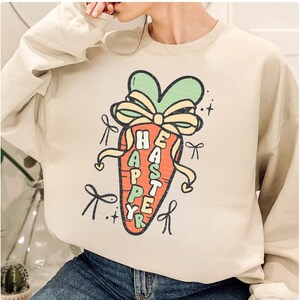 May include: A beige sweatshirt with a cartoon carrot graphic. The carrot is decorated with a green bow and the text "Happy Easter" is written on the carrot.