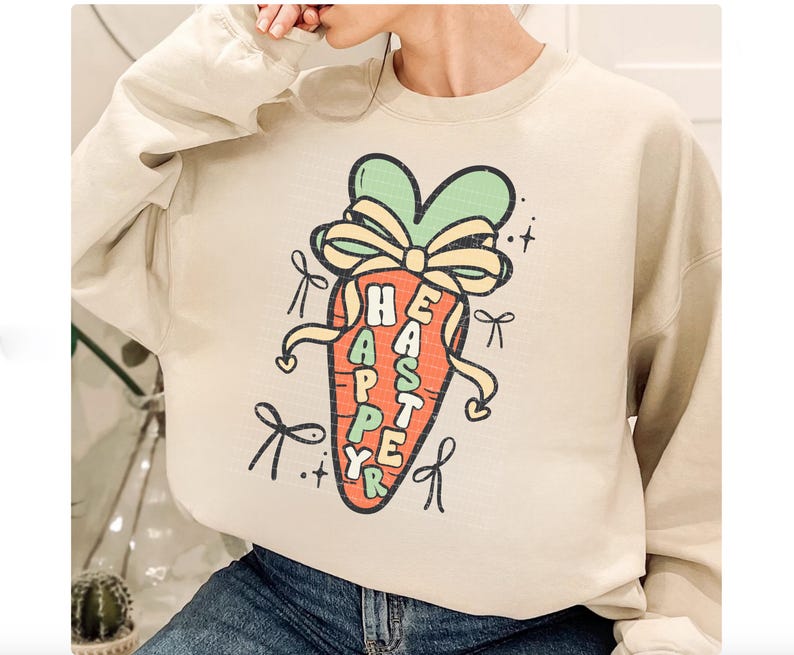 May include: A beige sweatshirt with a cartoon carrot graphic. The carrot is decorated with a green bow and the text "Happy Easter" is written on the carrot.