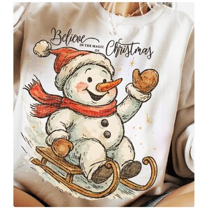 Op de afbeelding: Wit sweatshirt met een sneeuwpopontwerp. De sneeuwpop, met een rode muts en sjaal, zit op een houten slee. De tekst "Believe in the magic of Christmas" staat boven de sneeuwpop.