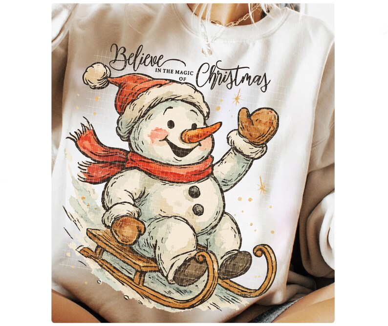 Op de afbeelding: Wit sweatshirt met een sneeuwpopontwerp. De sneeuwpop, met een rode muts en sjaal, zit op een houten slee. De tekst "Believe in the magic of Christmas" staat boven de sneeuwpop.