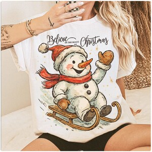 Op de afbeelding: Wit T-shirt met een vrolijke sneeuwman met een rode sjaal en hoed, op een houten slee. De tekst "Believe in the magic of Christmas" staat boven de sneeuwman. Een feestelijk thema.