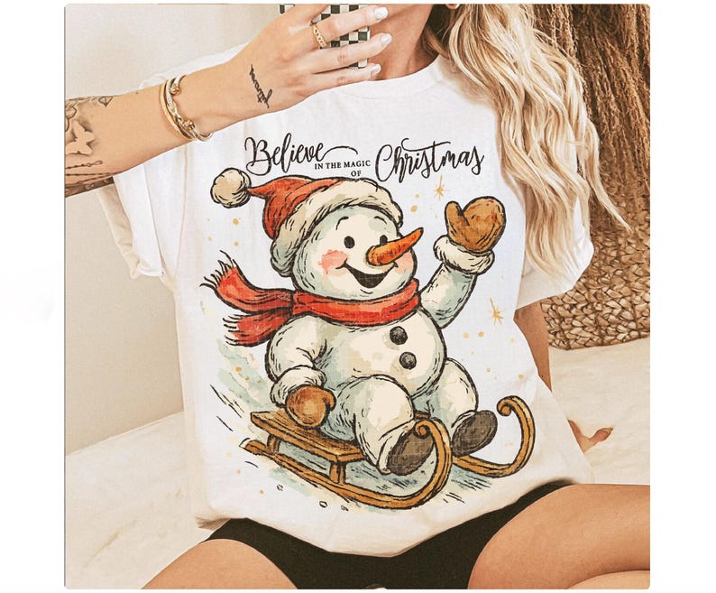 Op de afbeelding: Wit T-shirt met een vrolijke sneeuwman met een rode sjaal en hoed, op een houten slee. De tekst "Believe in the magic of Christmas" staat boven de sneeuwman. Een feestelijk thema.