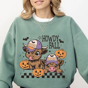 Puede incluir: Una sudadera de color verde azulado con un gr&aacute;fico de dos vacas de las tierras altas de dibujos animados que llevan sombreros y sostienen calabazas. El texto "Howdy Fall" est&aacute; impreso sobre las vacas.