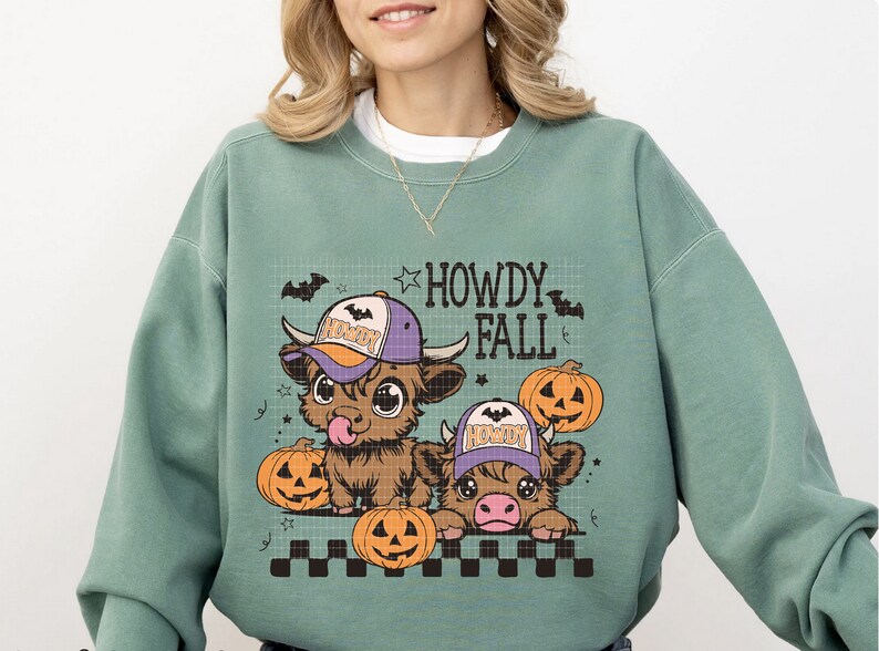 Puede incluir: Una sudadera de color verde azulado con un gr&aacute;fico de dos vacas de las tierras altas de dibujos animados que llevan sombreros y sostienen calabazas. El texto "Howdy Fall" est&aacute; impreso sobre las vacas.