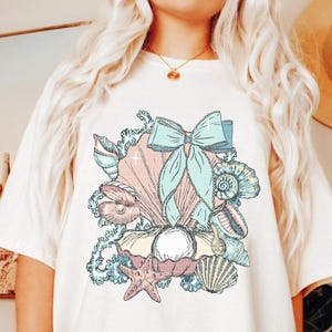 Puede incluir: Una camiseta blanca con una ilustraci&oacute;n colorida de conchas marinas, una perla, una estrella de mar y un lazo azul. La ilustraci&oacute;n es de estilo vintage.