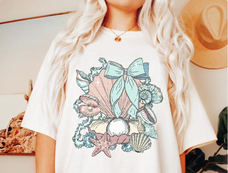 Puede incluir: Una camiseta blanca con una ilustraci&oacute;n colorida de conchas marinas, una perla, una estrella de mar y un lazo azul. La ilustraci&oacute;n es de estilo vintage.