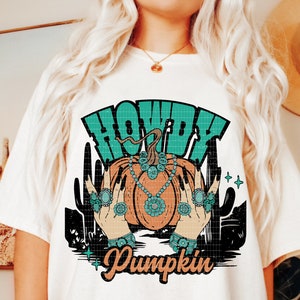 May include: A white t-shirt with a turquoise and orange graphic design featuring a pumpkin with a turquoise necklace and the text "Howdy Pumpkin".
