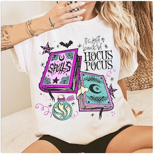 May include: White t-shirt with a colourful graphic featuring two spell books, a potion bottle, stars, and a bat. The text on the shirt reads "It's just a bunch of Hocus Pocus."