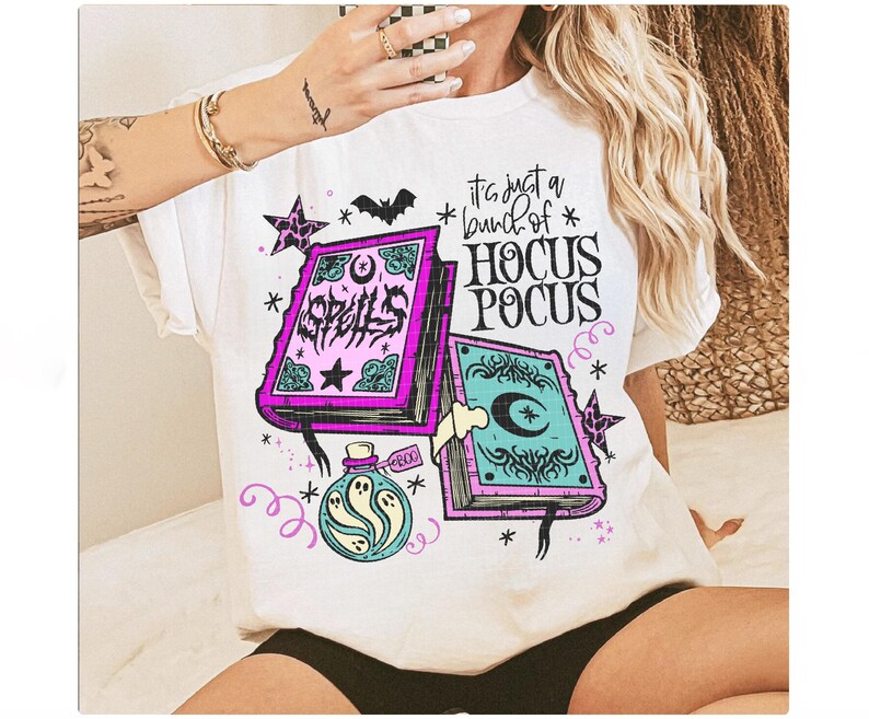 May include: White t-shirt with a colourful graphic featuring two spell books, a potion bottle, stars, and a bat. The text on the shirt reads "It's just a bunch of Hocus Pocus."