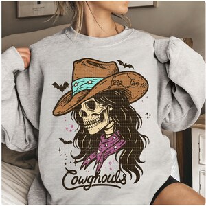 May include: A light gray sweatshirt featuring a graphic of a skull wearing a cowboy hat with the text "Long Live" and "Cowghouls". The hat is brown with a turquoise band and stars. The skull has long brown hair and a purple bandana.