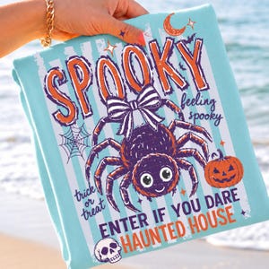 May include: A light blue t-shirt featuring a graphic print of a spider with a black and white striped bow, a pumpkin, a skull, and a spiderweb. The text reads SPOOKY, feeling spooky, trick or treat, and ENTER IF YOU DARE HAUNTED HOUSE in orange and purple.