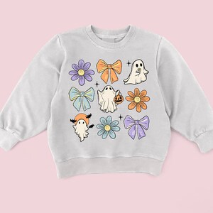 May include: A light gray sweatshirt with a Halloween theme. The sweatshirt features a variety of cute illustrations, including ghosts, flowers, and bows in shades of purple, blue, orange, and yellow.