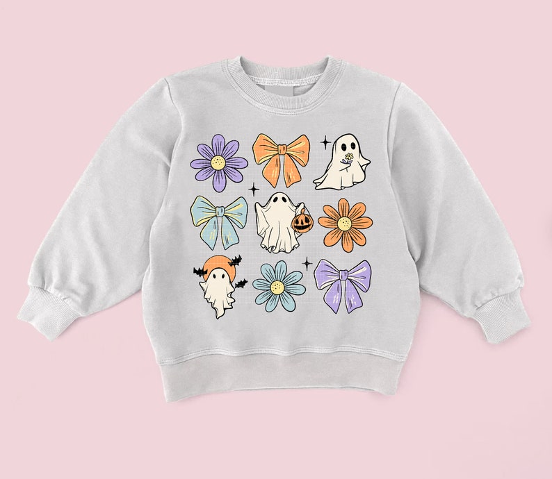May include: A light gray sweatshirt with a Halloween theme. The sweatshirt features a variety of cute illustrations, including ghosts, flowers, and bows in shades of purple, blue, orange, and yellow.