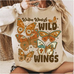 以下が含まれることがあります： ベージュのスウェットシャツ。蝶のグラフィックと「Western Winds & Wild Wings」の文字入り。蝶はオレンジ、茶色、ターコイズ色で、花柄と幾何学模様が施されています。スウェットシャツはクルーネックと長袖です。