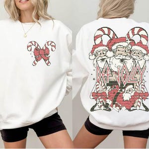 May include: White sweatshirt with a red and white candy cane design on the left side and a Christmas-themed graphic on the right side. The graphic features three Santa Clauses with the text "Eat Jolly" in a bold font.