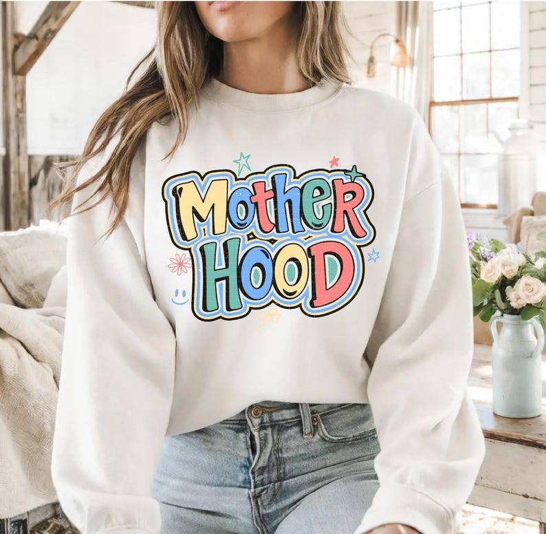May include: A cream-coloured sweatshirt featuring the word "Motherhood" in a colourful, playful font. The text is outlined in black and uses pastel colours such as yellow, blue, green, and pink. Stars and a smiling flower accent the design.